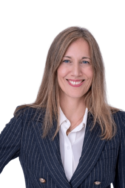 Angela Gamba, a woman with long, straight, light brown hair, smiles at the camera. She is wearing a white blouse and a dark pinstripe blazer with gold buttons. The background is transparent.