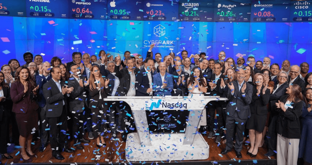 A large group of people celebrate at the Nasdaq podium, surrounded by falling blue confetti, with stock market screens displaying company logos and prices in the background.