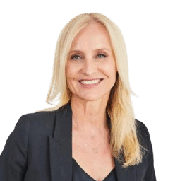 Dr. Eveline Steinberger, a smiling woman with long blonde hair, is wearing a dark suit and black top. She has one hand in her pocket and stands confidently against a plain white background.